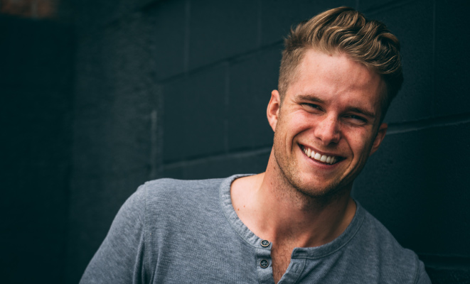 Blonde young man smiles with remineralized teeth while wearing a gray shirt against a teal wall