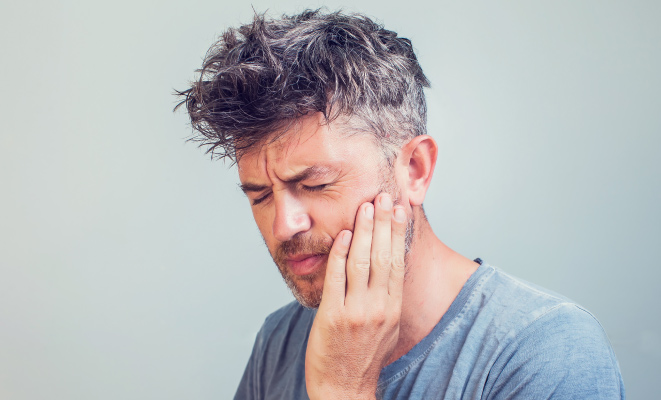 A middle-aged man wearing a gray shirt touches his cheek and winces due to phantom tooth pain