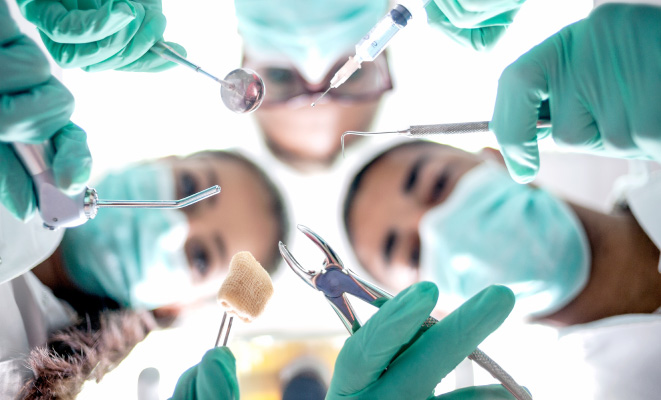 Looking up into 3 dentists performing oral surgery while wearing turquoise scrubs and holding dental tools and masks