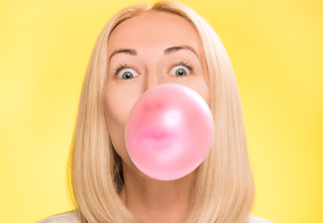 Blonde woman blows a pink gum bubble that contains xylitol against a yellow background