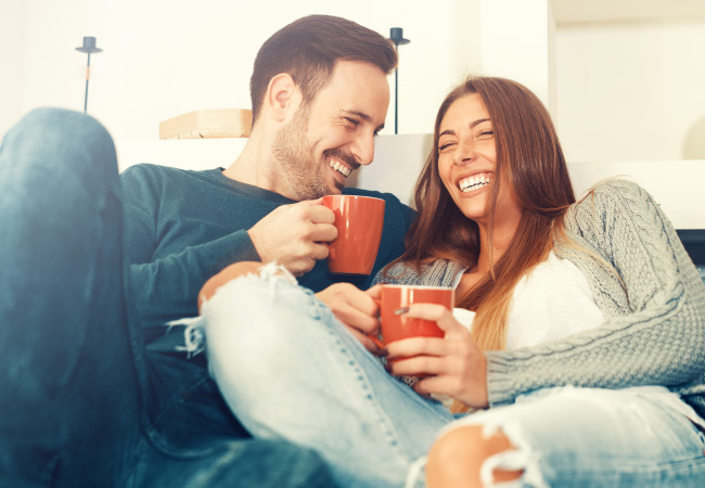 After receiving teeth whitening, a man and woman cuddle on the couch and smile with their orange mugs
