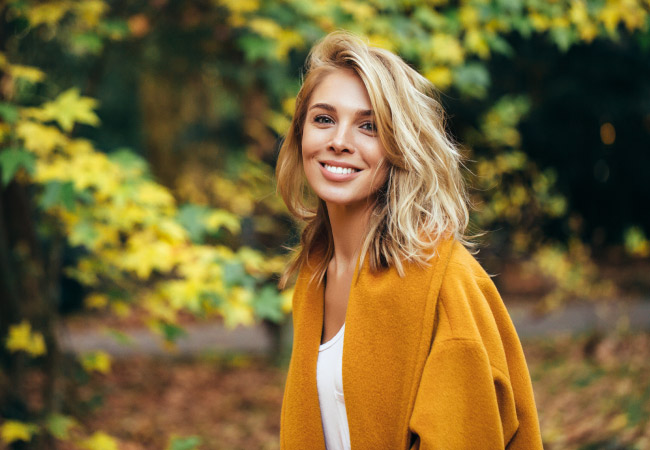 Blonde woman with dental veneers smiles outside by fall leaves wearing a mustard yellow cardigan